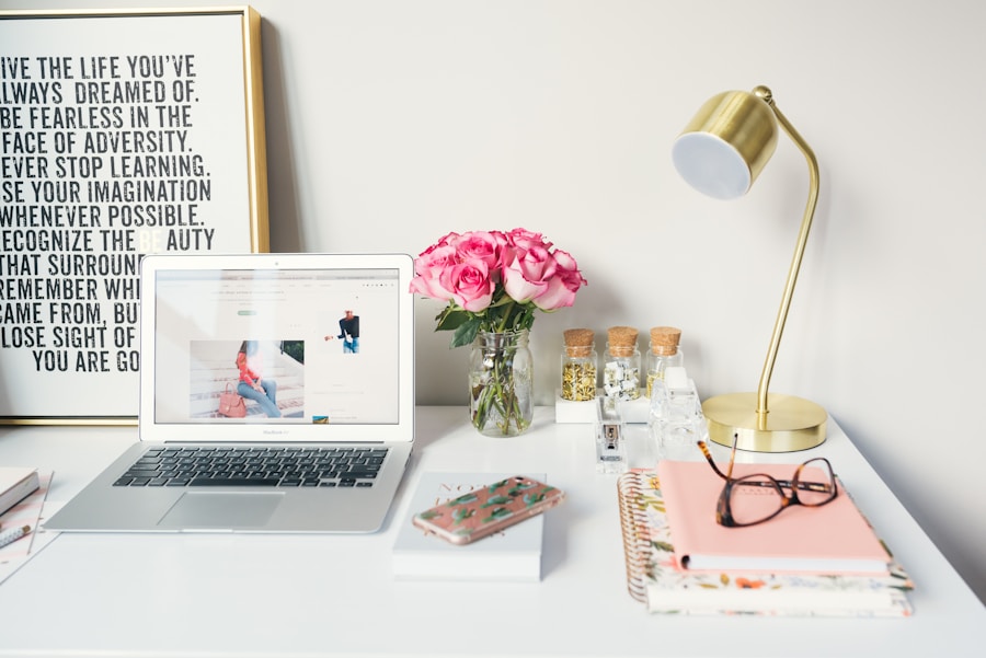 Laptop, pink roses, gold desk lamp, notebooks, and glasses on a white desk beside a framed quote poster